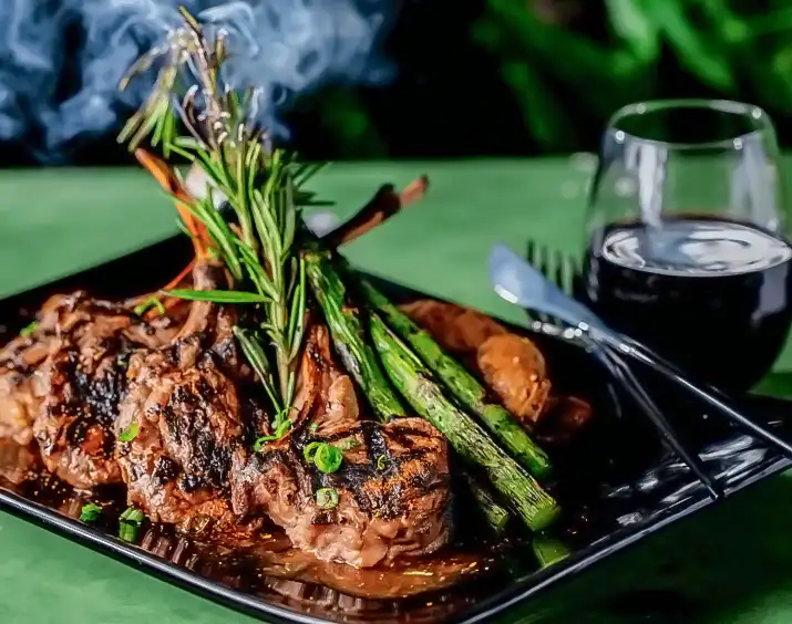 Miso marinated lamb chops with grilled asparagus and rosemary garnish, served on a black plate with a glass of red wine — Casa Sensei, Fort Lauderdale
