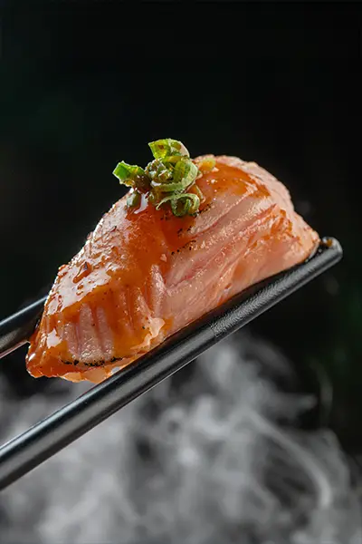 Glazed salmon nigiri held by black chopsticks over rising steam