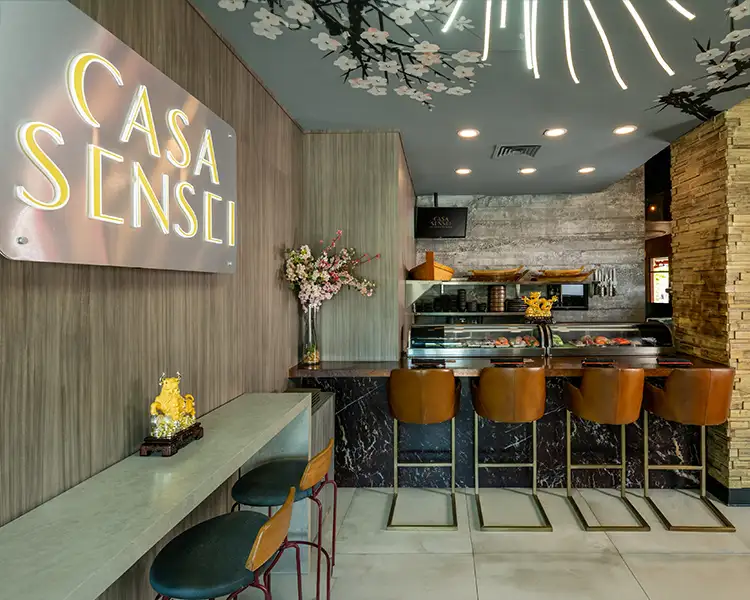 Interior of Casa Sensei restaurant featuring a dark marble sushi bar with leather counter stools, illuminated neon logo, and cherry blossom ceiling decor