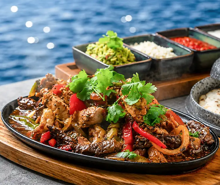 Sizzling chicken fajitas with grilled peppers, onions, fresh cilantro, tortillas, and colorful toppings beside the water.
